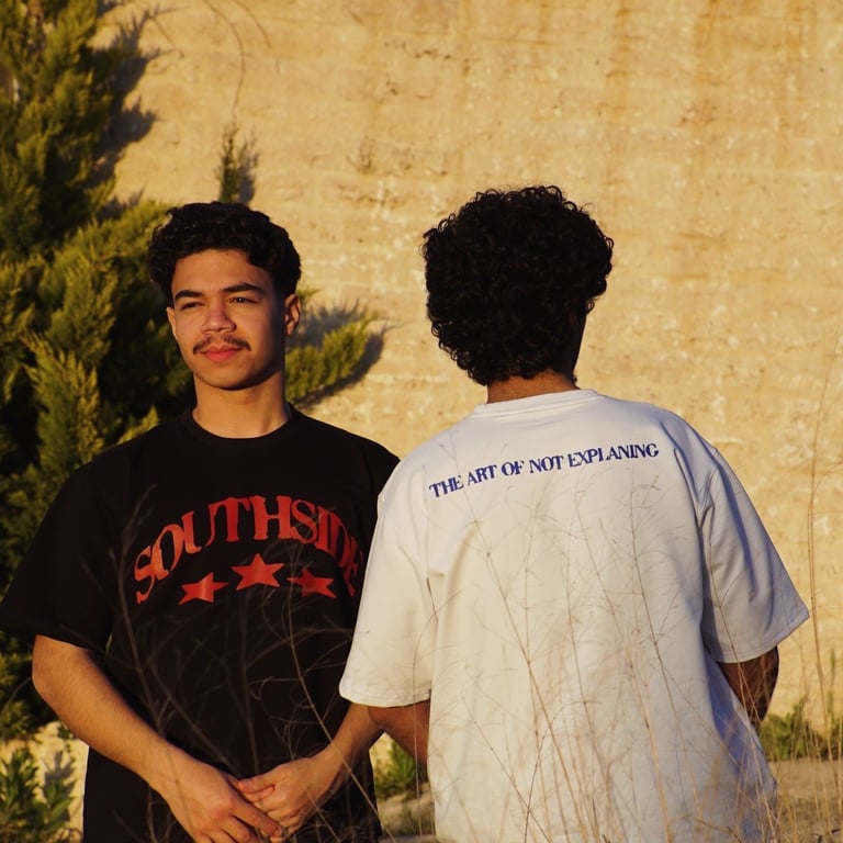 Two young men standing against a textured beige wall with green ivy, wearing graphic t-shirts